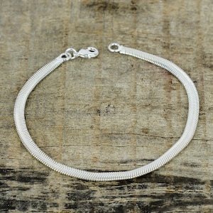 Pode incluir: Uma pulseira prateada em espinha de peixe com fecho de lagosta. A pulseira é feita de pequenas ligações interligadas, criando um design flexível em forma de cobra. A pulseira é exibida em uma superfície de madeira.