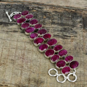 Puede incluir: Una pulsera de plata con piedras preciosas ovaladas de color rojo intenso. Cada piedra preciosa está engastada en un bisel de plata y conectada por eslabones de plata. La pulsera tiene un cierre de palanca y descansa sobre una superficie de madera.