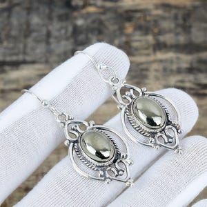 May include: A pair of silver-toned dangle earrings. Each earring features an oval, metallic gray stone set within an ornate, filigree design. The earrings have a hook closure.