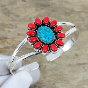 May include: A silver cuff bracelet with a central oval turquoise stone, encircled by red oval stones. The bracelet has a floral design. The background is a neutral colour.