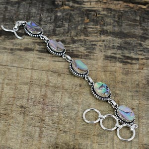 Puede incluir: Una pulsera de plata con piedras ovaladas de concha de abulón. Las piedras iridiscentes están engastadas en un bisel con cuentas y conectadas por eslabones de plata. Cierre de gancho.