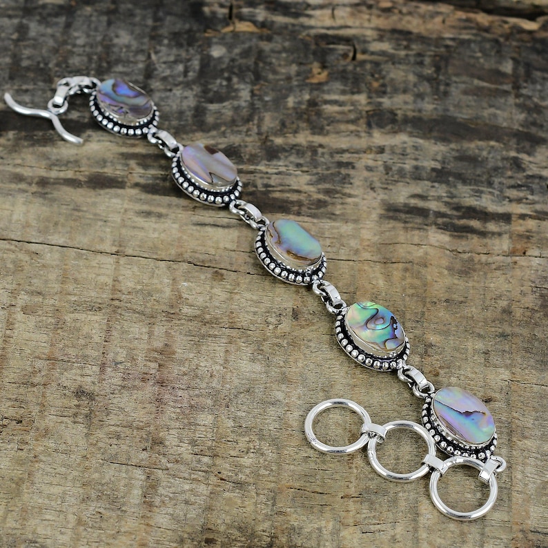 K&ouml;nnte beinhalten: Ein silbernes Armband mit ovalen Abalone-Muschelsteinen. Die irisierenden Steine sind in einer Perlenfassung gefasst und durch Silberglieder verbunden. Das Armband hat einen Hakenverschluss.