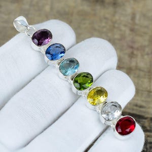 May include: A silver pendant with seven oval gemstones in a row. The gemstones are in various colours, including purple, blue, green, yellow, and red. The pendant is held by a gloved hand.