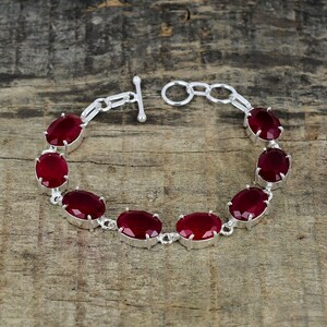 May include: A silver bracelet with a toggle clasp features seven oval-shaped red gemstones.