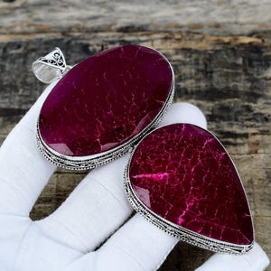 May include: Two large, oval and teardrop-shaped pendants with a deep magenta gemstone set in ornate silver-colored metal. The gemstones have a cracked, textured appearance. The pendants are displayed on a wooden surface.