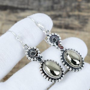 May include: A pair of silver-coloured drop earrings. Each earring has an oval, metallic stone set in a beaded frame. The design includes a decorative swirl and a hook for the ear.