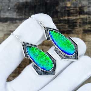 May include: A pair of dangle earrings featuring elongated, teardrop-shaped iridescent stones with vibrant green and blue color shifts. The stones are set in oxidized silver frames with a textured, dark finish and attached to simple silver ear wires.