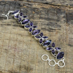 Pode incluir: Uma pulseira de prata com pedras preciosas roxas profundas com corte marquesa, dispostas em um design floral. A pulseira, com fecho de alternância, é exibida em uma superfície de madeira.