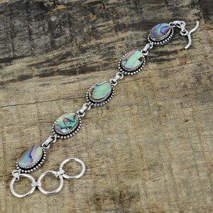 K&ouml;nnte beinhalten: Ein silbernes Armband mit ovalen Abalone-Muschelschmucksteinen. Jeder Stein ist in einer Perlenfassung gefasst und durch Silberglieder verbunden. Die schillernden Muscheln zeigen Farbt&ouml;ne von Gr&uuml;n, Blau und Lila. Das Armband hat einen Hakenverschluss und drei Verbindungsringe.