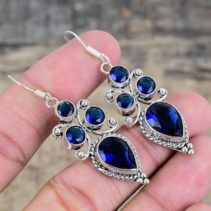 Pode incluir: Um par de brincos prateados com um design floral. Cada brinco apresenta múltiplas pedras preciosas redondas e em forma de gota, azul escuro. Os brincos são segurados por uma mão.