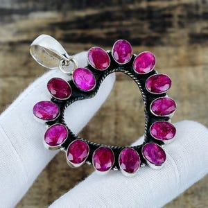 Puede incluir: Un colgante en forma de corazón que presenta una serie de piedras preciosas de color rosa rojizo vibrante, de corte ovalado, engastadas a lo largo de un marco de plata trenzada. La pieza incluye una reasa de plata pulida en la parte superior.