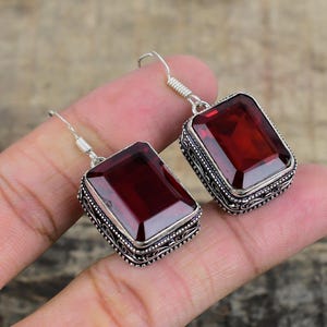 Op de afbeelding: Een paar zilveren oorbellen met grote, rechthoekige, dieprode edelstenen. De edelstenen zijn gezet in sierlijke, gedetailleerde zilveren monturen. De oorbellen hebben een haaksluiting.