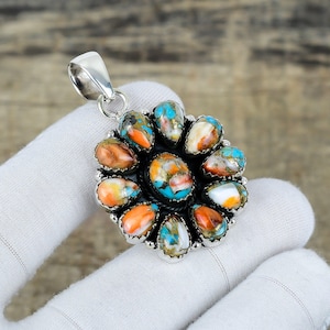 Peut inclure: Un pendentif en argent orné d'un groupe de pierres précieuses colorées. Le pendentif présente une pierre turquoise centrale entourée de pierres orange, blanches et bleues. Les pierres précieuses sont serties dans un sertissage en argent avec une boucle pour une chaîne.