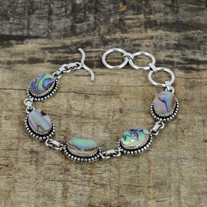 K&ouml;nnte beinhalten: Ein silbernes Armband mit ovalen Abalone-Muschelschmucksteinen. Die irisierenden Steine sind in einer Perlenfassung gefasst und durch Silberglieder verbunden. Das Armband hat einen Hakenverschluss.