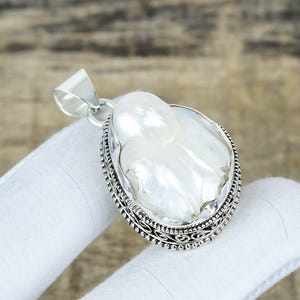 Pode incluir: Pingente de prata com uma grande pérola branca de formato irregular. A pérola está emoldurada em um quadro oval com detalhes intrincados em prata nas bordas. O pingente tem uma argola simples para uma corrente.