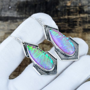 May include: A pair of dangle earrings featuring teardrop-shaped iridescent stones with vibrant pink, green, and blue flashes. The stones are set in textured, oxidized silver frames with a geometric, angular design and silver hook ear wires.