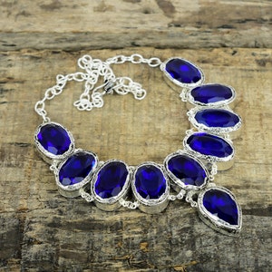 May include: A silver necklace with multiple oval and teardrop-shaped, deep blue gemstones. The gemstones are set in a silver metal frame and connected by a delicate chain. The necklace is displayed on a rustic wooden surface.