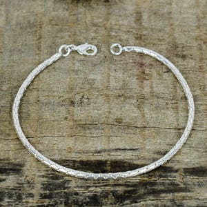 Puede incluir: Una pulsera plateada con un diseño texturizado en forma de serpiente. La pulsera es circular y presenta un cierre de mosquetón. La joya se exhibe sobre una superficie de madera.