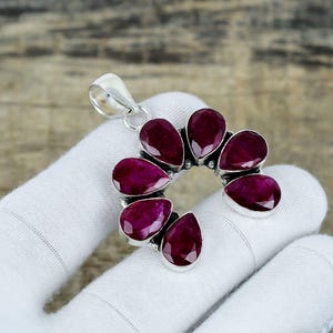 May include: A silver pendant with seven teardrop-shaped, deep red gemstones set in a semi-circle. The gemstones are mounted in a silver frame, with a silver loop for a chain. The pendant is shown on a white surface.