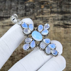 Puede incluir: Colgante plateado en forma de cruz, adornado con un óvalo central y piedras preciosas en forma de lágrima, azul claro y blanco. El colgante tiene una argolla texturizada para una cadena.