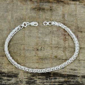 Könnte beinhalten: Ein silbernes Armband mit einem geflochtenen Kettendesign. Das Armband besteht aus einem glänzenden, hellfarbigen Metall und verfügt über einen Karabinerverschluss. Das Armband wird auf einer Holzoberfläche präsentiert.