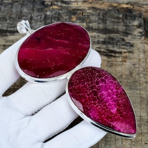 Könnte beinhalten: Zwei Edelsteinanhänger mit silberfarbenem Rand. Einer ist oval, der andere tropfenförmig. Beide haben einen tiefmagentafarbenen Stein mit strukturierter Oberfläche. Der ovale Anhänger hat eine silberne Schlaufe.