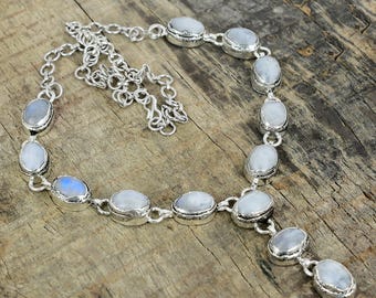 Puede incluir: Un collar de plata con cabujones ovalados de piedra lunar iridiscente. Las piedras están engastadas en biseles individuales y conectadas por una delicada cadena. El collar se exhibe sobre una superficie de madera.