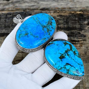 Puede incluir: Dos colgantes de turquesa con engastes de metal plateado. Los colgantes son de color azul brillante con vetas verdes oscuras y marrones. Uno es ovalado y el otro tiene forma de lágrima. Los colgantes se muestran sobre una superficie blanca.