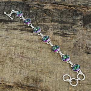 Puede incluir: Un brazalete de plata con cierre de palanca presenta seis piedras redondas e iridiscentes. Las piedras preciosas tienen un arcoíris de colores, incluyendo verde, azul, morado y rosa.