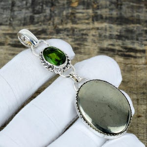 Puede incluir: Un colgante de plata con una piedra preciosa ovalada de color verde oliva y una piedra grande ovalada de color gris metálico. El colgante está conectado por eslabones de plata y tiene un lazo en la parte superior para una cadena.