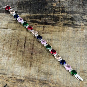 May include: A silver-toned tennis bracelet featuring a repeating pattern of round, multi-coloured gemstones including red, light pink, dark blue, and green, accented by small clear stones set between each larger coloured stone in a metal link chain.