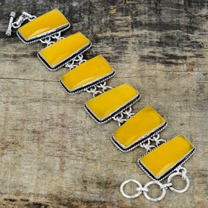 Puede incluir: Una pulsera de plata con piedras preciosas rectangulares amarillas. Las piedras están delineadas en negro y engastadas en un marco de plata. La pulsera tiene un cierre de palanca y se muestra sobre una superficie de madera.