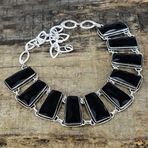 Puede incluir: Un collar plateado con grandes piedras negras rectangulares. Las piedras están engarzadas en un marco plateado y unidas por una delicada cadena. El collar se exhibe sobre una superficie de madera.