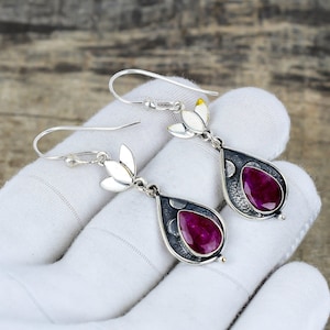 May include: A pair of silver earrings with a teardrop-shaped, dark gray setting. Each earring features a large, oval-cut, deep red gemstone. The earrings have a floral design at the top and are displayed on a white surface.