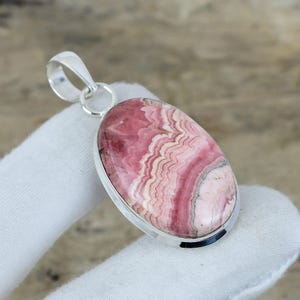 Könnte beinhalten: Ovaler Anhänger mit einem polierten Rhodochrosit-Edelstein, der rosa und weiße Bänder aufweist. Der Edelstein ist in einen silberfarbenen Metallrahmen mit einer Schlaufe für eine Kette gefasst.