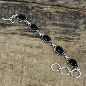 Peut inclure: Un bracelet en argent orné de pierres précieuses noires ovales. Le bracelet présente une bordure perlée décorative autour de chaque pierre et une fermeture à fermoir. Le bijou est présenté sur une surface en bois.