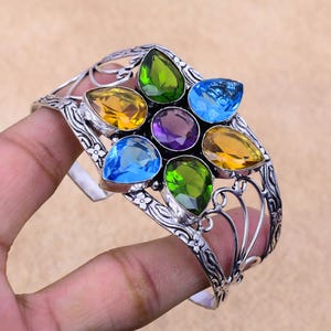 May include: A silver bangle bracelet with a floral design and seven faceted gemstones. The gemstones are in shades of green, blue, yellow, and purple.