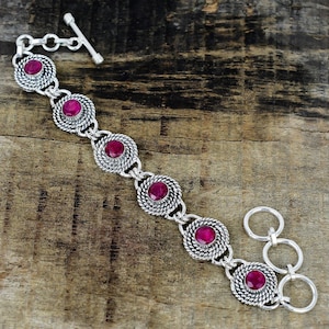 Op de afbeelding: Een zilveren armband met zes cirkelvormige schakels, elk voorzien van een ronde, gefacetteerde, donkerroze edelsteen. De schakels zijn verbonden door zilveren ringen en de armband wordt gesloten met een T-sluiting en drie extra ringen.