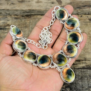 May include: A silver-toned necklace featuring large, round, polished shells with a mix of colors including green, brown, and cream. The shells are set in individual bezels and connected by a chain. The necklace is displayed on a wooden surface.