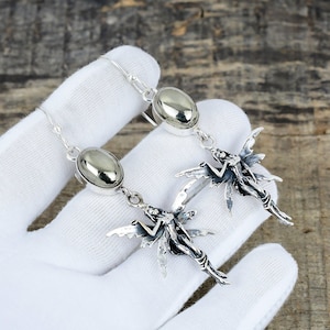 Può includere: Orecchini pendenti color argento con design a fata. Ogni orecchino presenta una pietra ovale lucida e un ciondolo a forma di fata dettagliato con ali spiegate. Gli orecchini sono esposti su sfondo bianco.