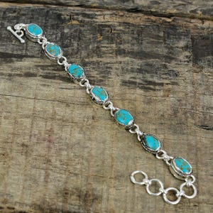 May include: A silver bracelet featuring oval turquoise stones. The stones are set in a silver bezel and linked together with silver chain. The bracelet has a toggle clasp closure. The turquoise stones have a vibrant blue-green color.