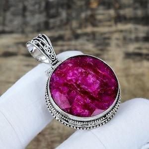 以下が含まれることがあります： 銀色の金属フレームにセットされた、大きな鮮やかなピンク色の宝石が付いた丸いペンダント。 宝石は、より暗い静脈を持つテクスチャの外観をしています。 ペンダントには、チェーンを取り付けるための装飾的なベイルが付いています。