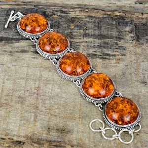Könnte beinhalten: Ein silbernes Armband mit fünf großen, ovalen Bernsteinen. Die Bernsteine haben eine warme, durchscheinende orange Farbe und sind in verzierten Silberfassungen gefasst. Das Armband hat einen Knebelverschluss und wird auf einer Holzoberfläche präsentiert.