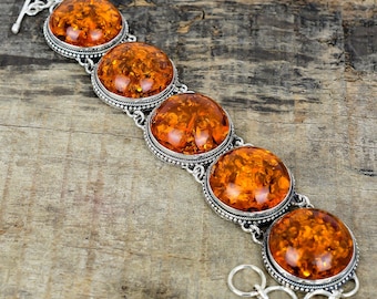 May include: A silver bracelet featuring five large, oval amber stones. The amber stones are a warm, translucent orange color, set in ornate silver bezels. The bracelet has a toggle clasp and is displayed on a wooden surface.