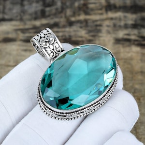 Puede incluir: Colgante ovalado con una gran gema azul claro facetada. La gema está engastada en un marco de metal plateado con intrincados adornos y una fianza decorativa. El colgante se muestra sobre un fondo blanco.