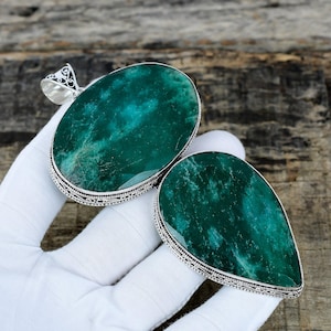 Puede incluir: Dos colgantes de piedras preciosas con engaste plateado. Uno es ovalado, el otro en forma de lágrima. Ambos presentan una piedra verde moteada. Los colgantes se muestran sobre un fondo de madera.