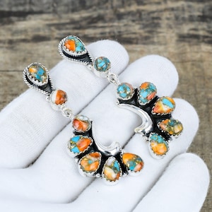 May include: Silver-toned earrings with a cluster of colorful stones. The stones feature a mix of orange, turquoise, and gold hues. The earrings have a unique, curved design, with a teardrop-shaped top and a series of stones along the curve.