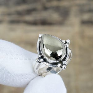 Puede incluir: Anillo de plata con una gran piedra de pirita pulida en forma de pera. La pirita tiene una superficie metálica y reflectante. El engaste del anillo tiene elementos decorativos en los laterales. El anillo se muestra sobre un fondo neutro.