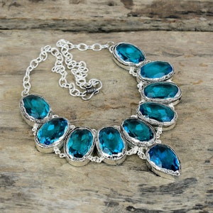 May include: A silver necklace with oval and teardrop-shaped teal gemstones. The gemstones are set in a silver bezel and linked by a silver chain. The necklace is shown on a wooden surface.