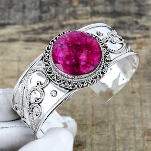 May include: A silver cuff bracelet with an oval, vibrant pink gemstone at the center. The bracelet features intricate scrollwork and a decorative border around the gemstone. The cuff is open-ended and has a polished finish.